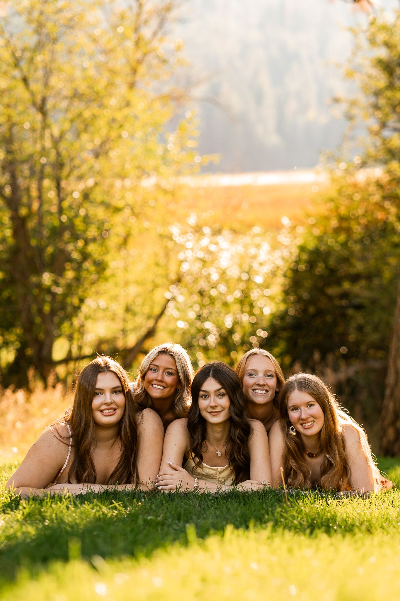 Five smiling young women lying on green grass outdoors in golden sunlight, wearing neutral tones, posing together for a group photo.