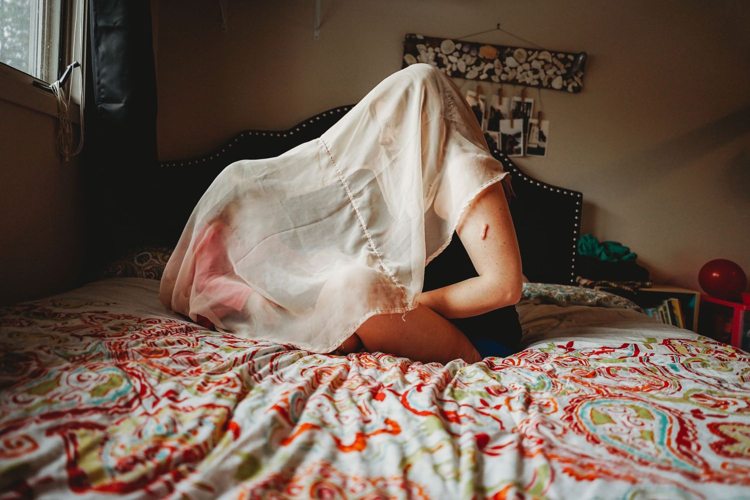 Person playfully hiding under a white sheet on a colorful patterned bed in a bedroom.