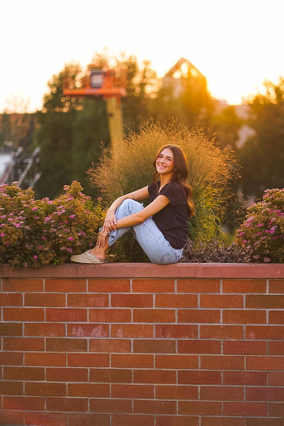 Smiling teen girl in jeans sitting cross-legged on brick wall with golden sunset background.