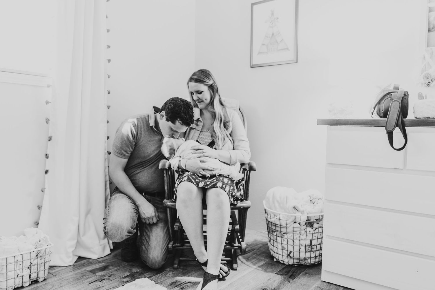 Black and white photo of parents with newborn in nursery chair surrounded by baby items.