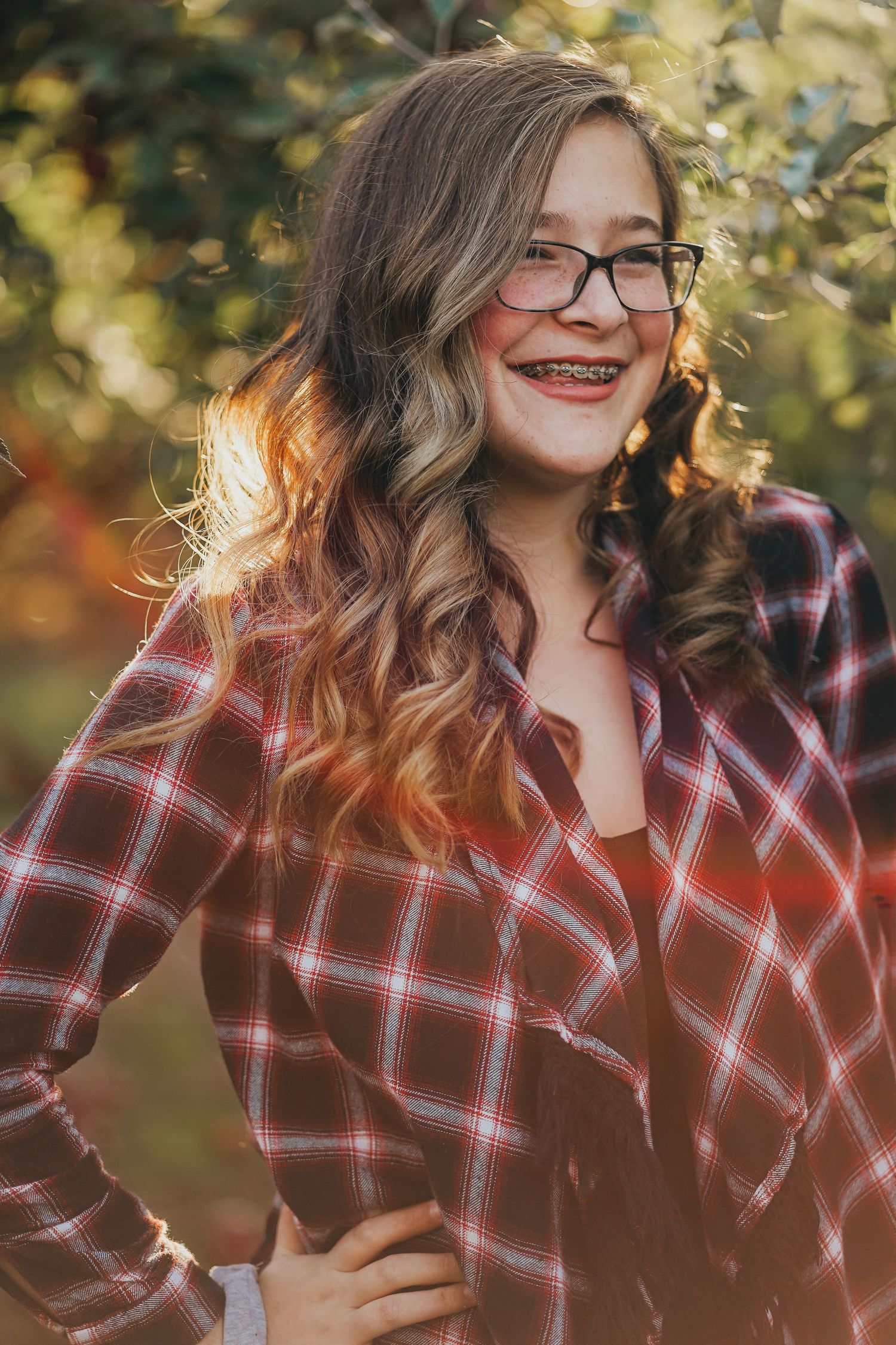 Smiling teenage girl with glasses and braces wearing a plaid shirt in a sunny outdoor portrait.
