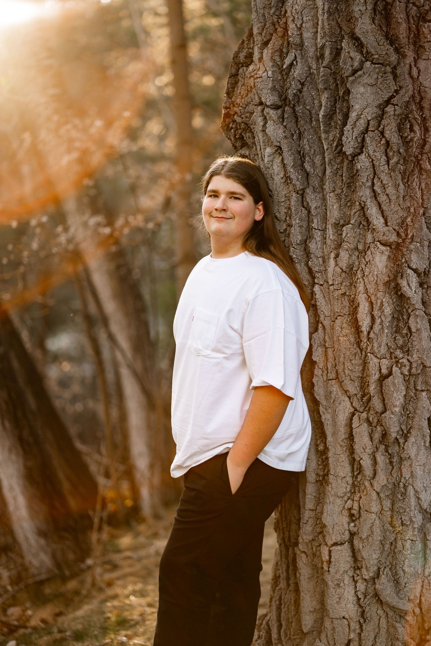 Teen with long hair leaning against large tree trunk in golden sunlight forest.