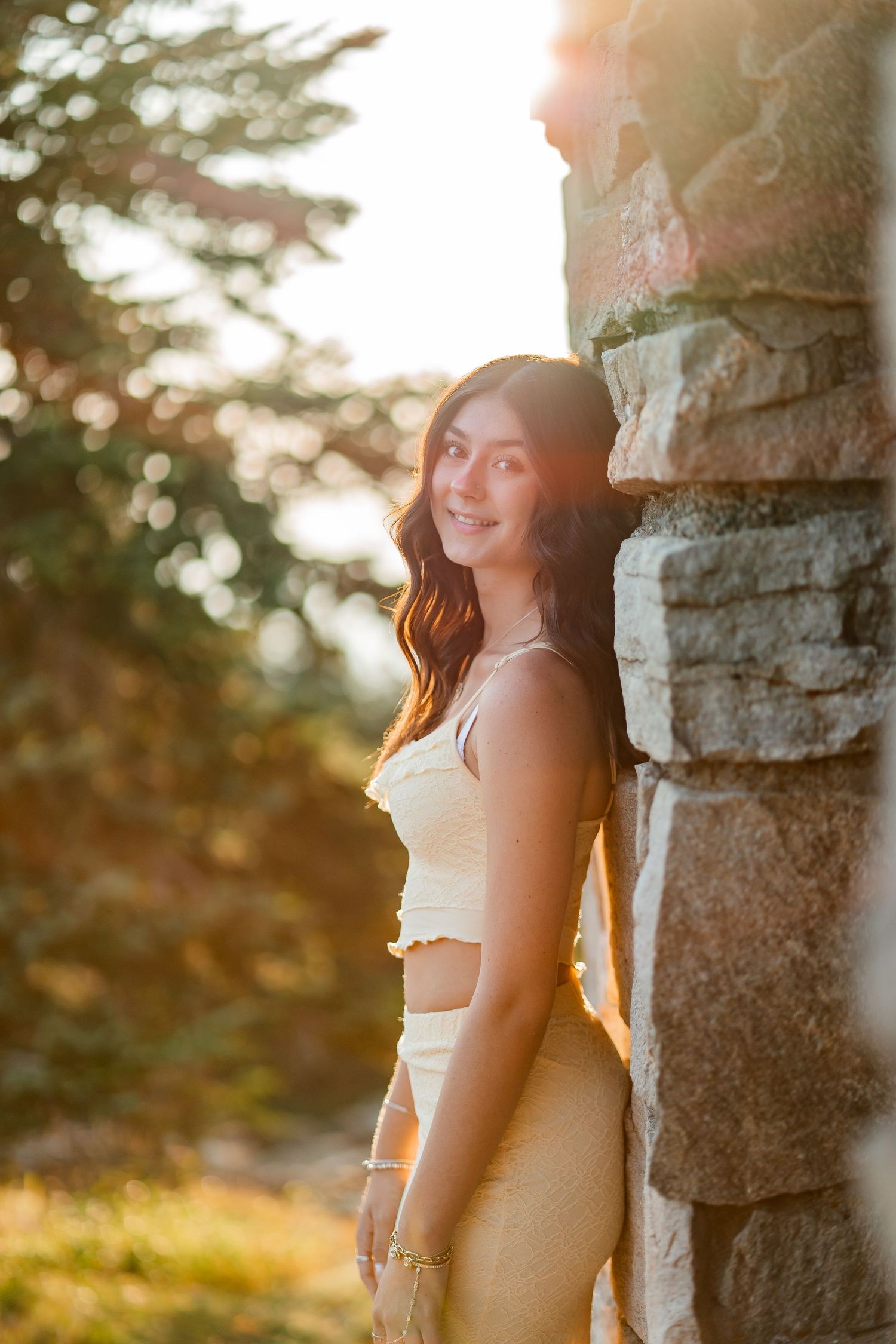 Young woman in cream two-piece outfit leaning against stone wall in golden sunlight.