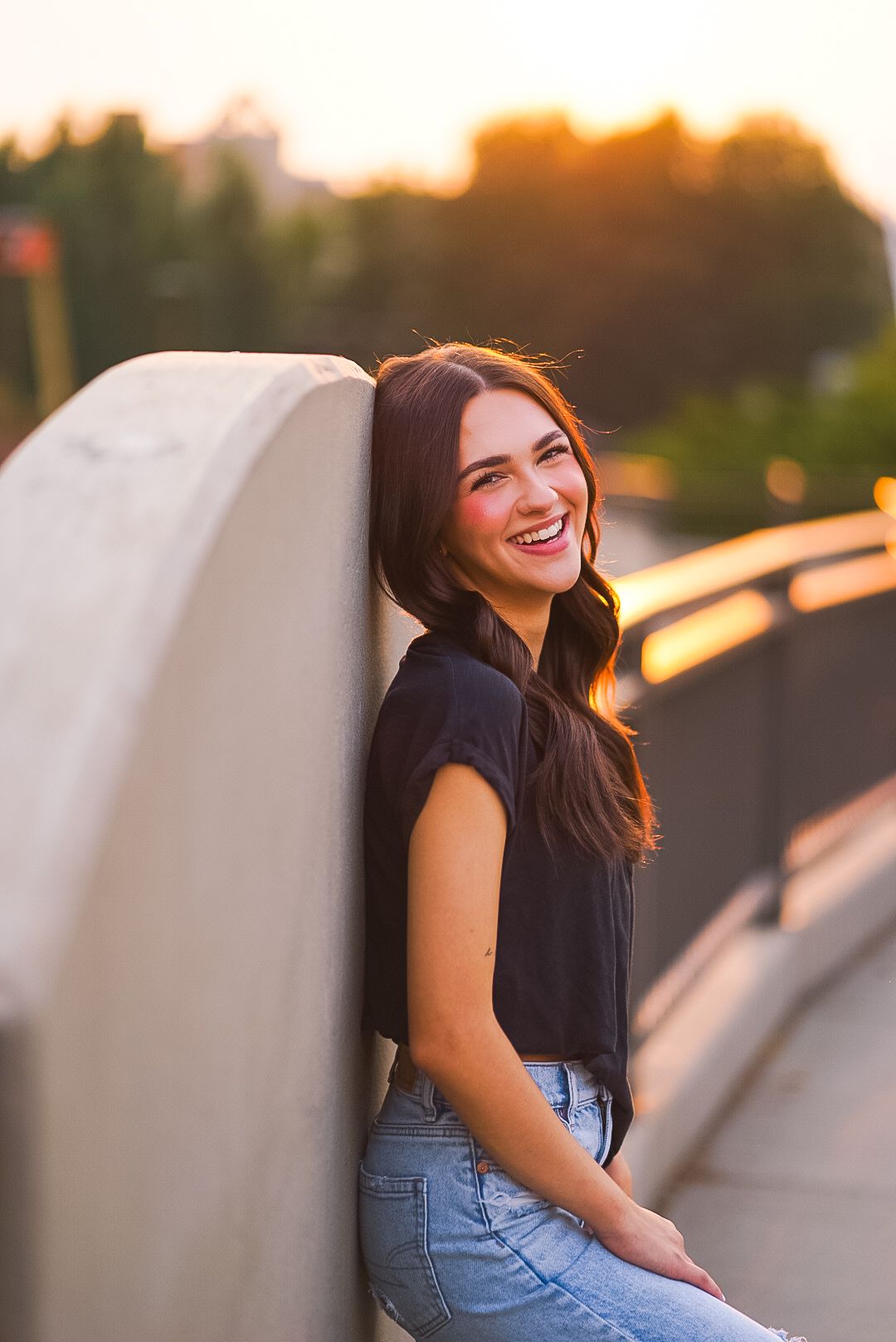 Joyful young woman in black tee and jeans leaning on wall laughing in golden sunset light.