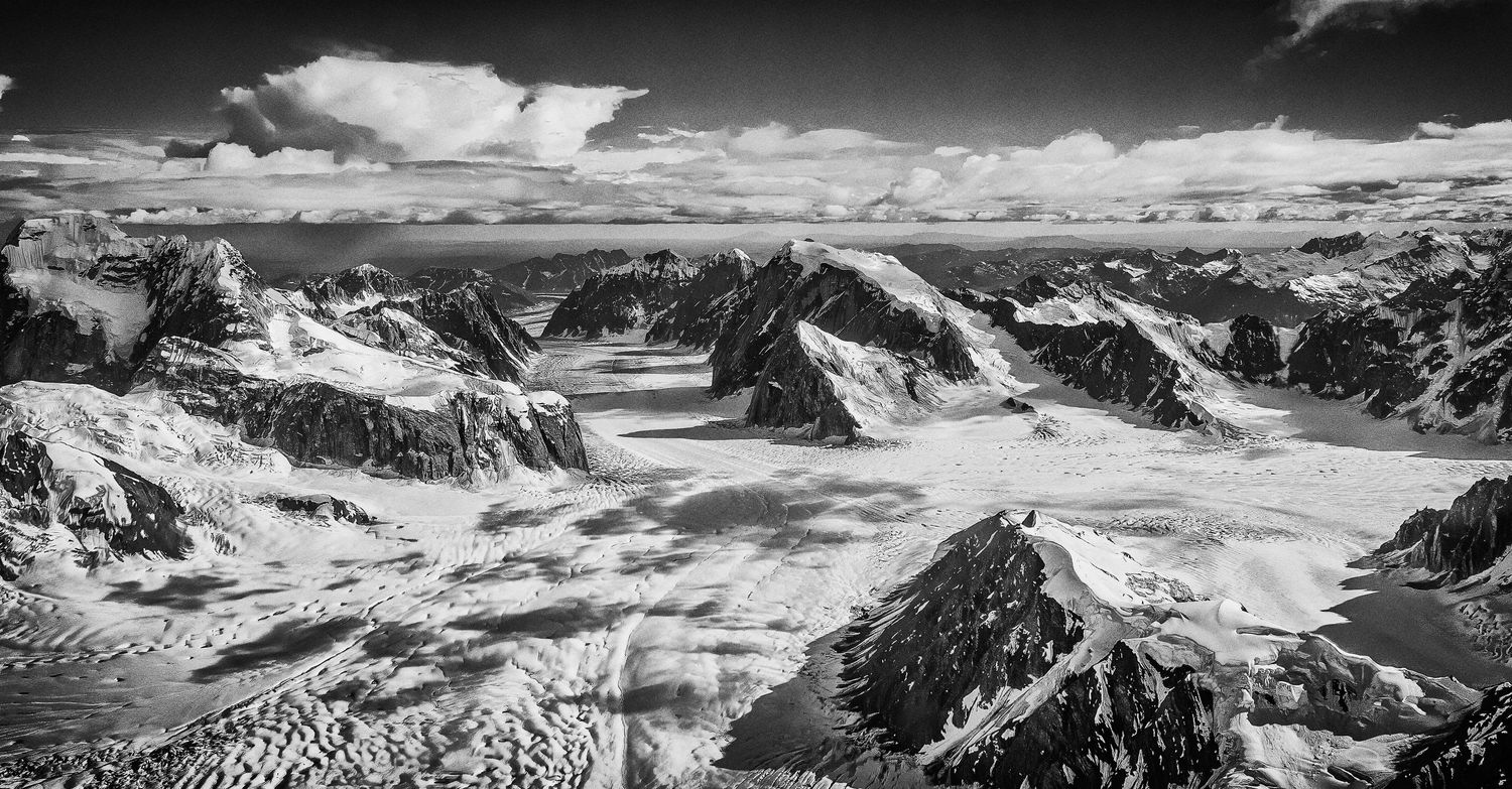 Black and white aerial panorama of vast glaciers and jagged mountain peaks under cloudy sky.