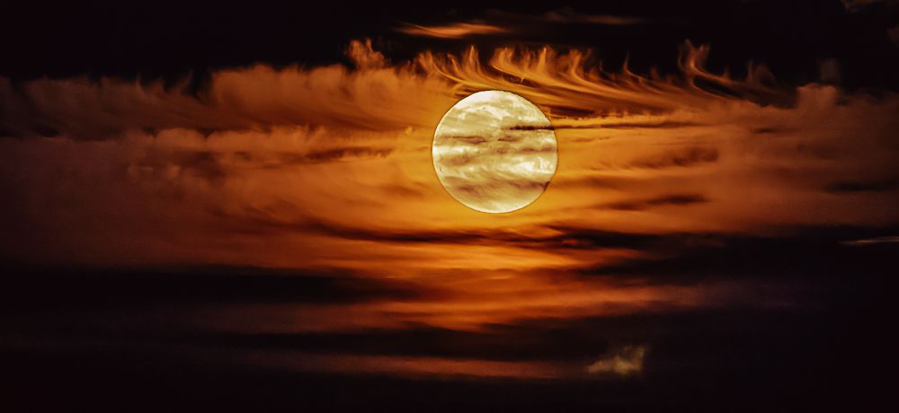 A dramatic full moon glows orange surrounded by dark swirling clouds in a night sky.