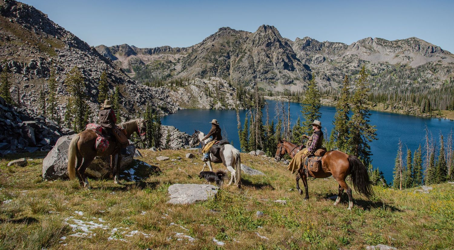 Three horseback riders traverse a scenic mountain trail beside a crystal-clear alpine lake.