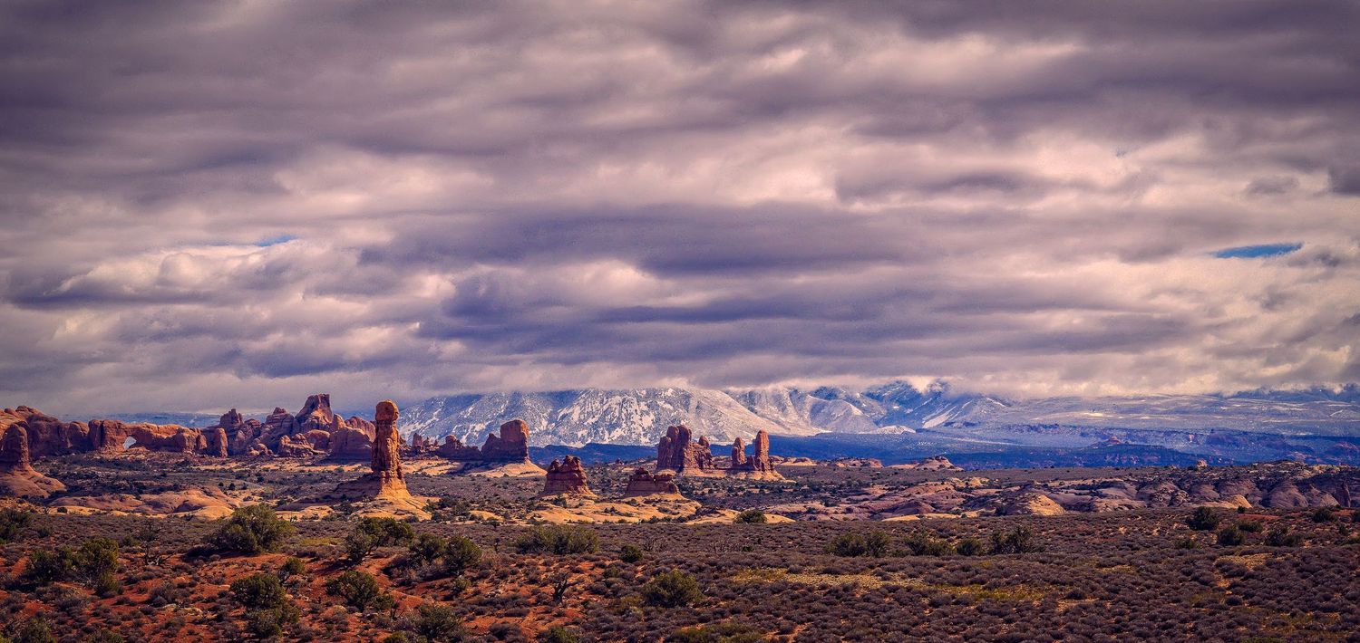 Panoramic view of Arches National Park with red rock formations and snow-capped mountains under dramatic clouds.