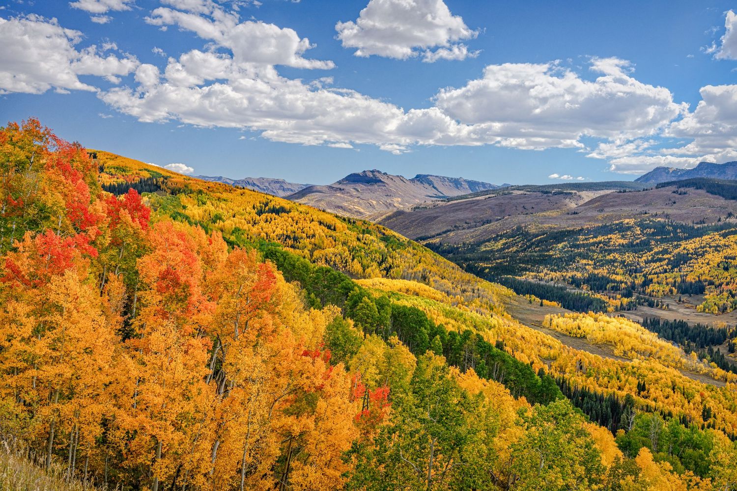 Vibrant autumn foliage with red, orange, and yellow trees covering Colorado mountain slopes under a partly cloudy blue sky.
