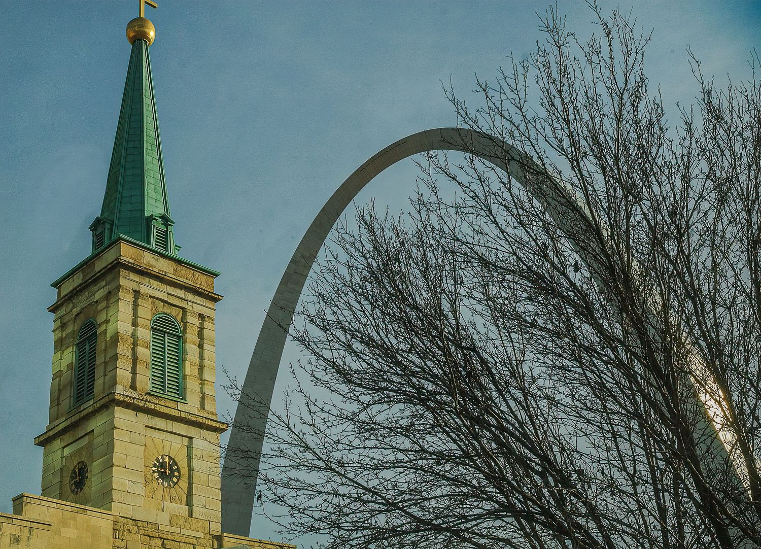 The iconic St. Louis Gateway Arch rises behind a historic church steeple and bare winter trees.
