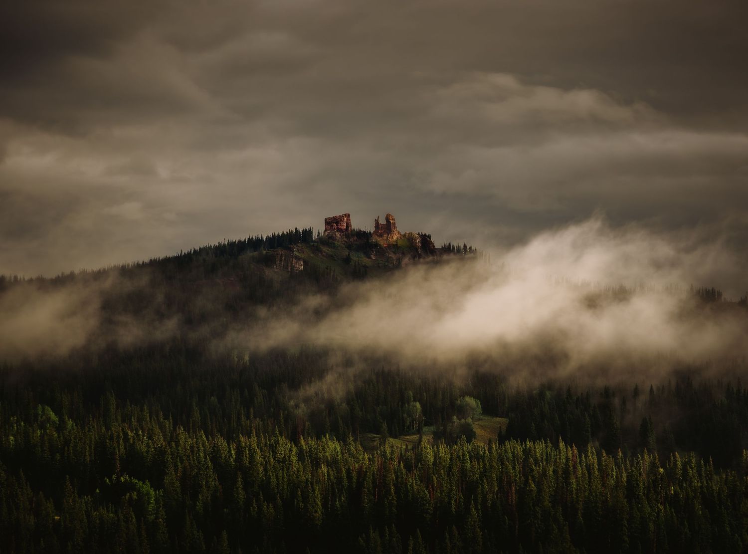 A medieval castle sits atop a forested hill shrouded in dramatic mist and dark storm clouds.