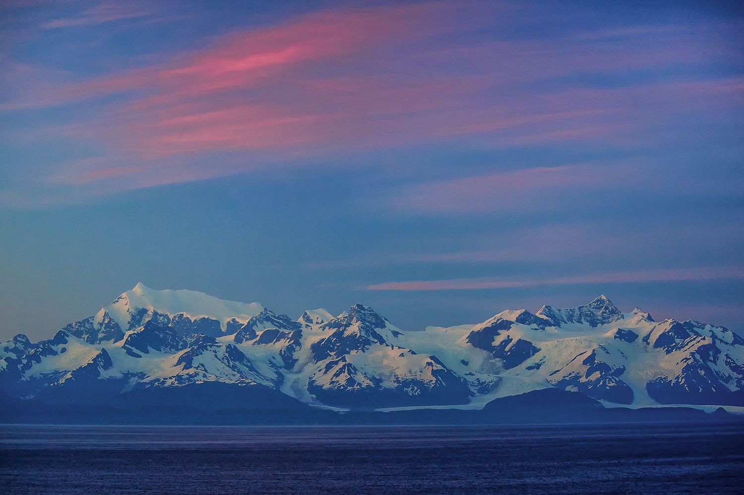 Snow-capped mountain range glows blue and pink under a dramatic sunset sky over calm Arctic waters.