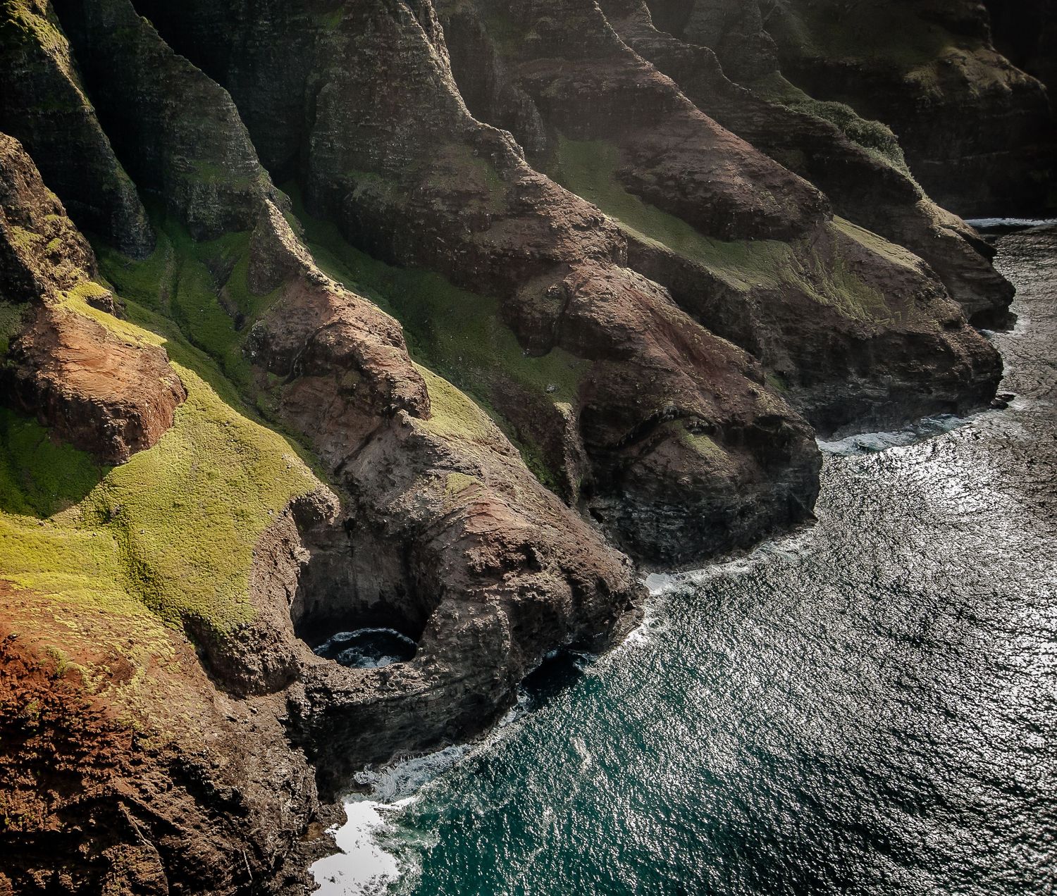 Dramatic aerial view of rugged Na Pali Coast sea cliffs with dark rock arches meeting turquoise ocean waves.