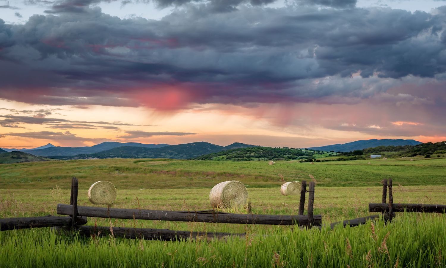 Hay bales on a green farm field at dramatic sunset with storm clouds over distant mountains.