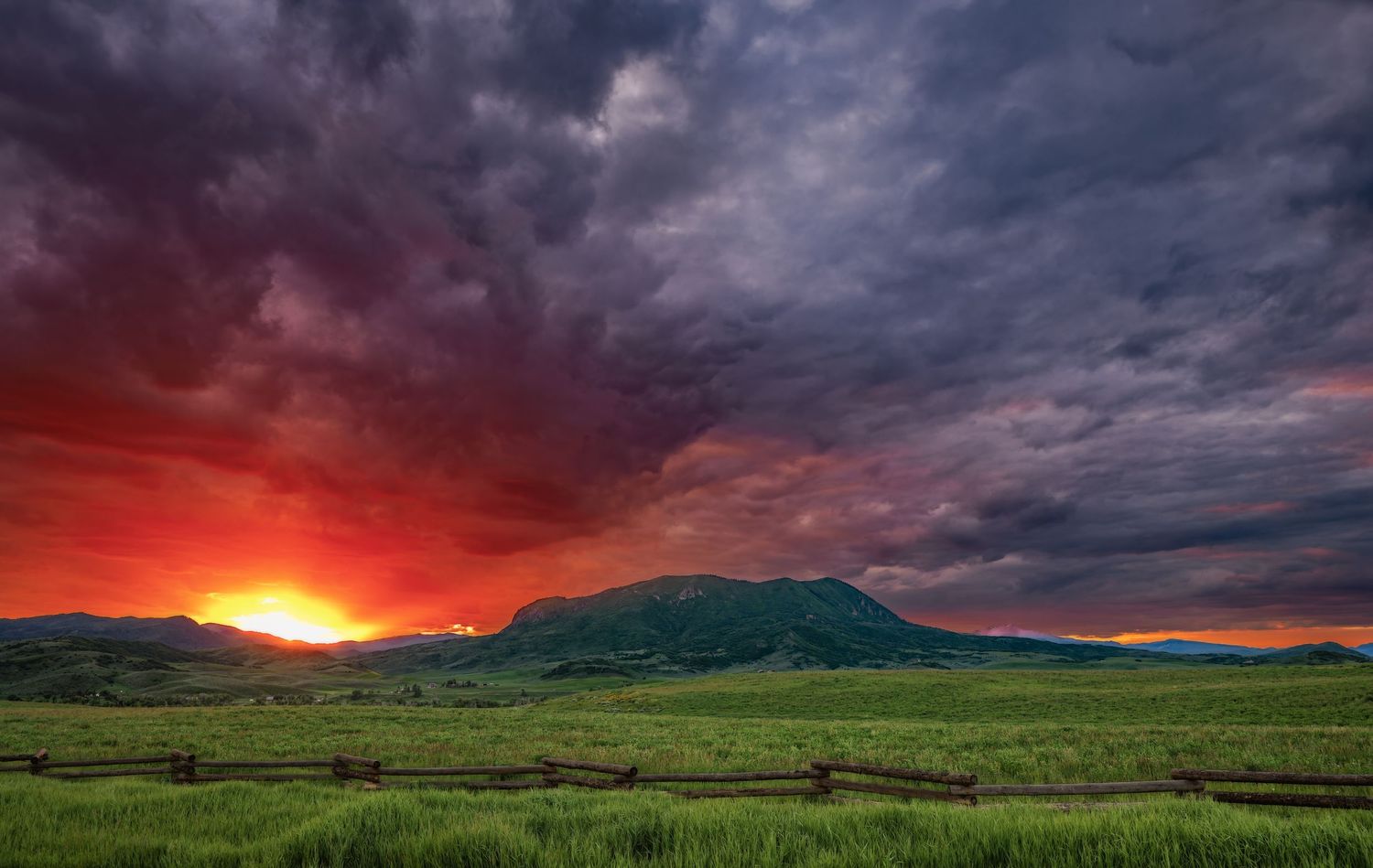 Dramatic fiery sunset with red and orange storm clouds over a green meadow and lone mountain silhouette.