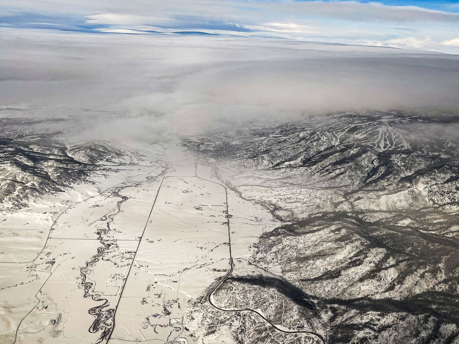 Aerial winter landscape showing snow-covered mountains, valleys, and a straight road under cloud cover.