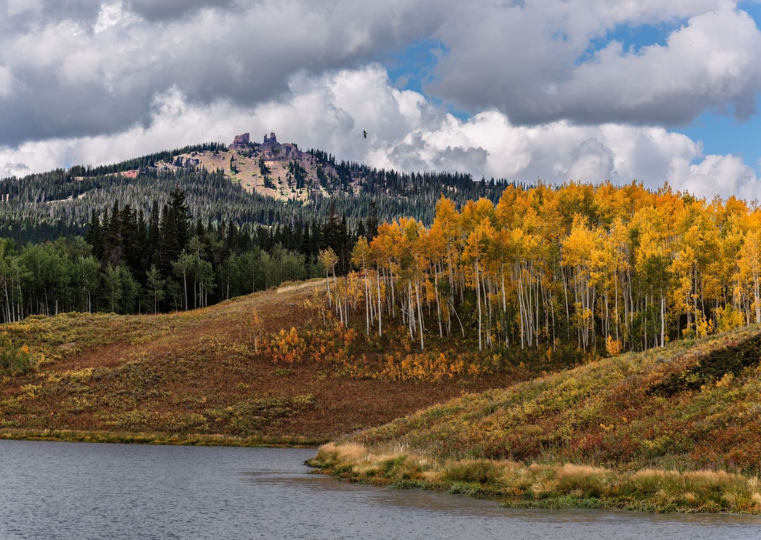 Golden aspen trees and rocky peaks reflect in a calm lake under dramatic autumn clouds.
