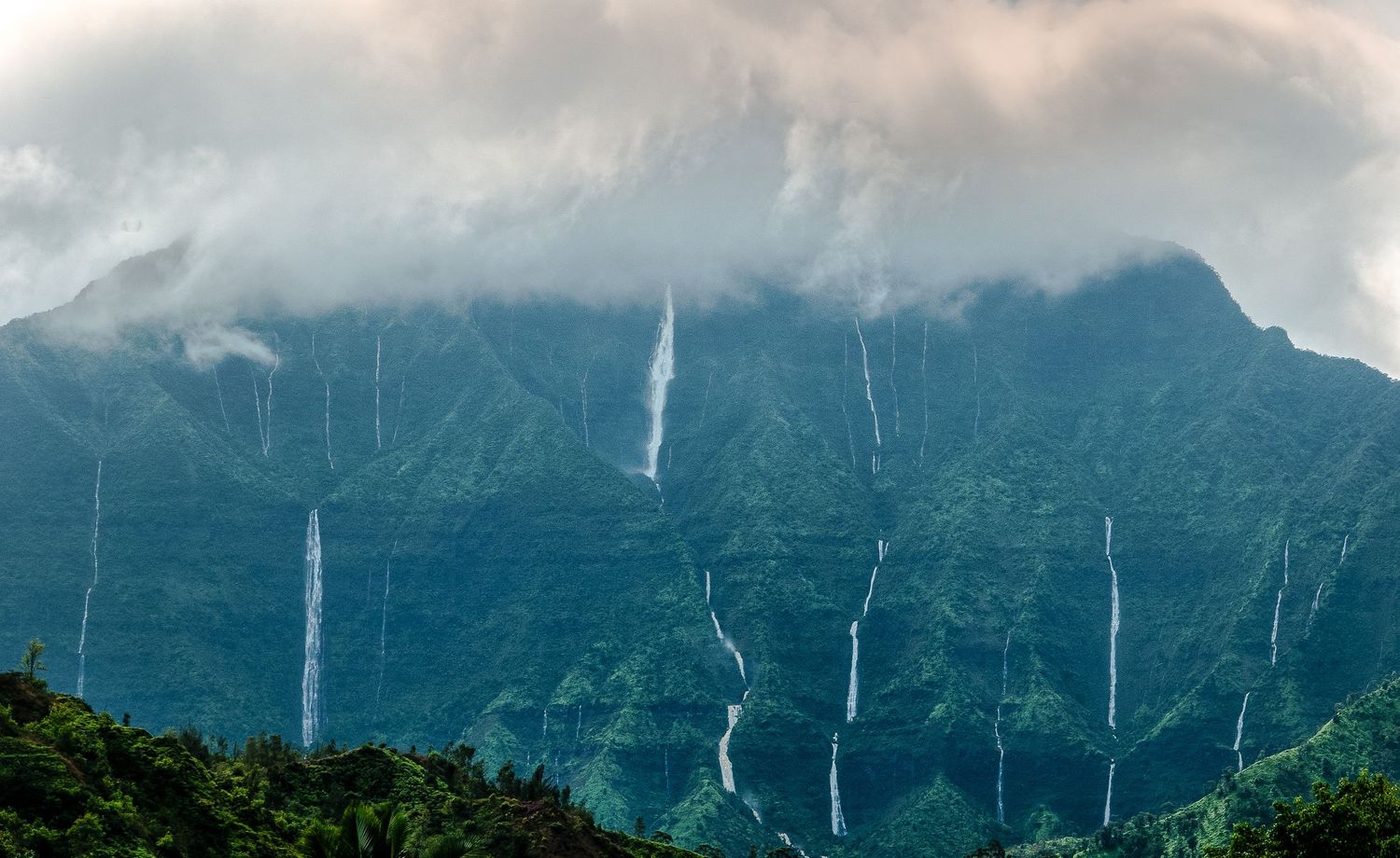 Dramatic multiple waterfalls cascade down a misty green mountain shrouded in clouds.