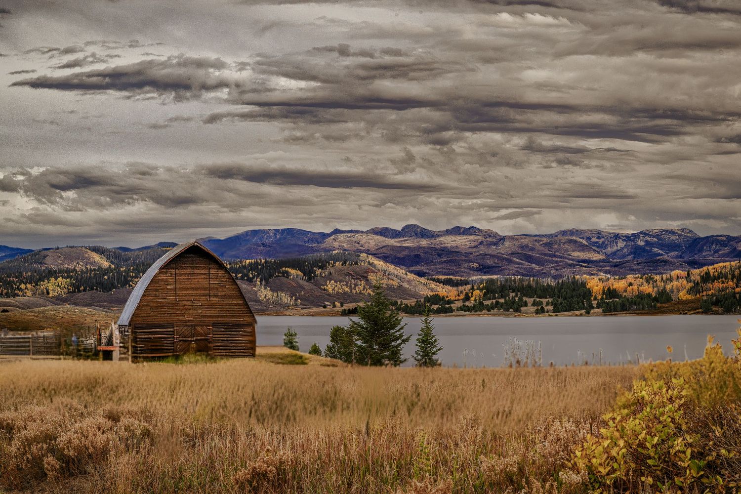 A rustic wooden barn sits beside a lake with mountains under stormy skies.