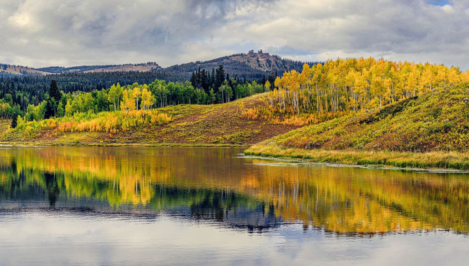 Calm lake reflecting golden autumn aspen trees and rolling hills in a serene mountain landscape.