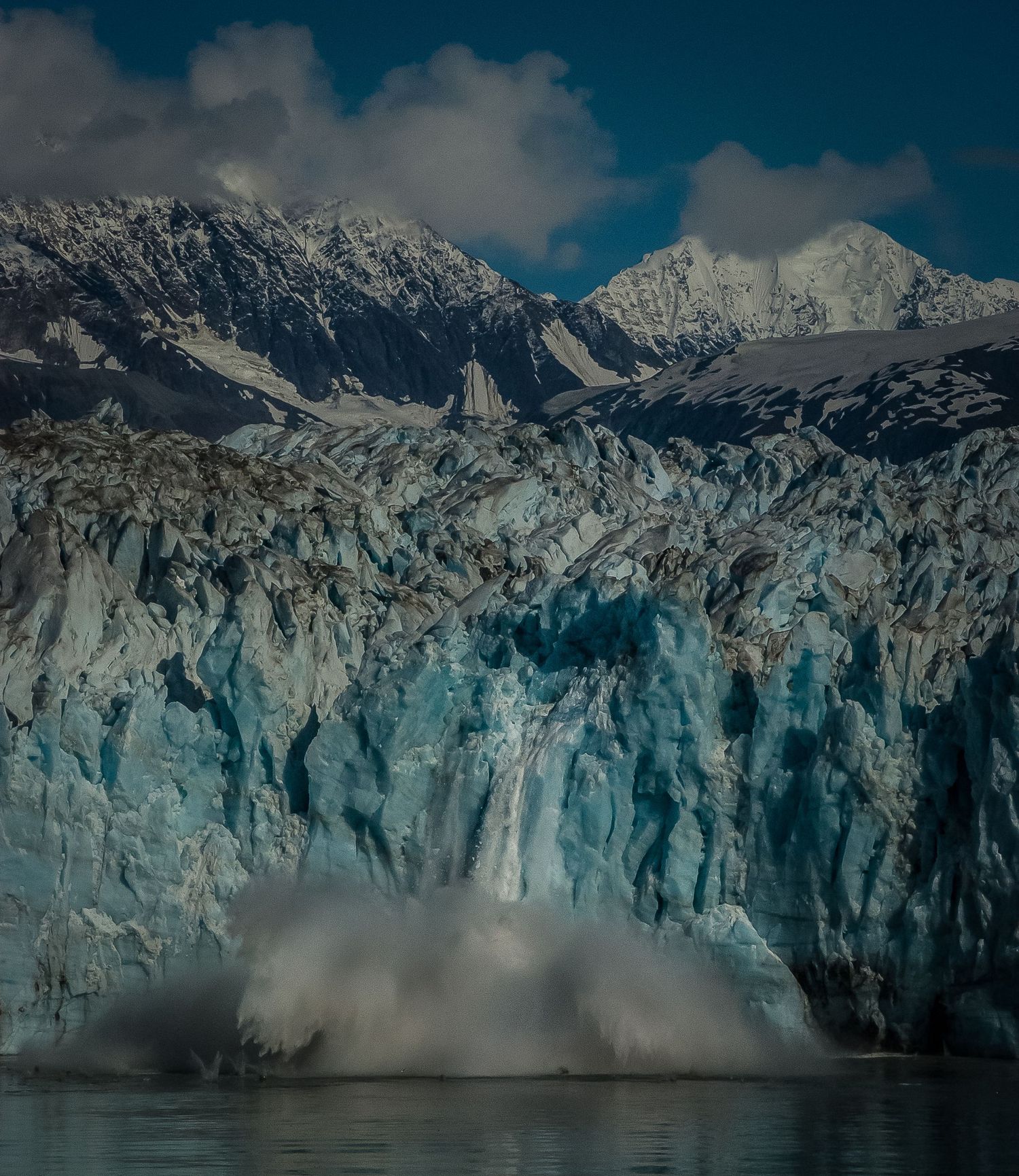 A massive glacier calving into the ocean with snow-capped mountains behind.
