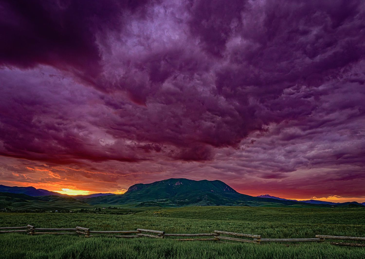 A dramatic purple and orange sunset storm sky glows over a green mountain meadow with a wooden fence.