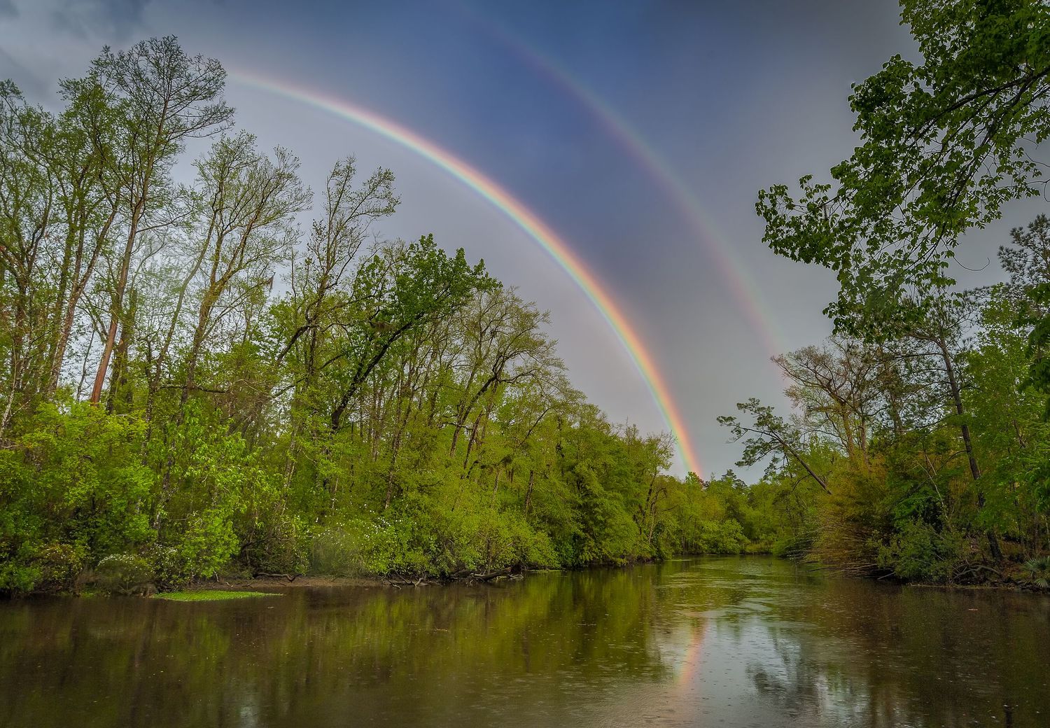 A stunning double rainbow arches over a calm river surrounded by lush green trees.