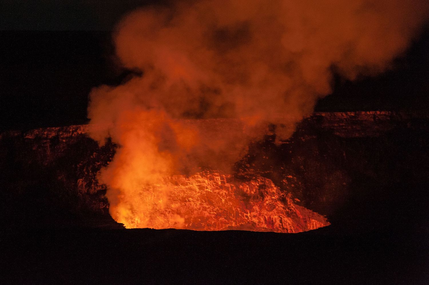 Glowing lava lake erupts with intense orange light and billowing smoke inside a volcanic crater at night.