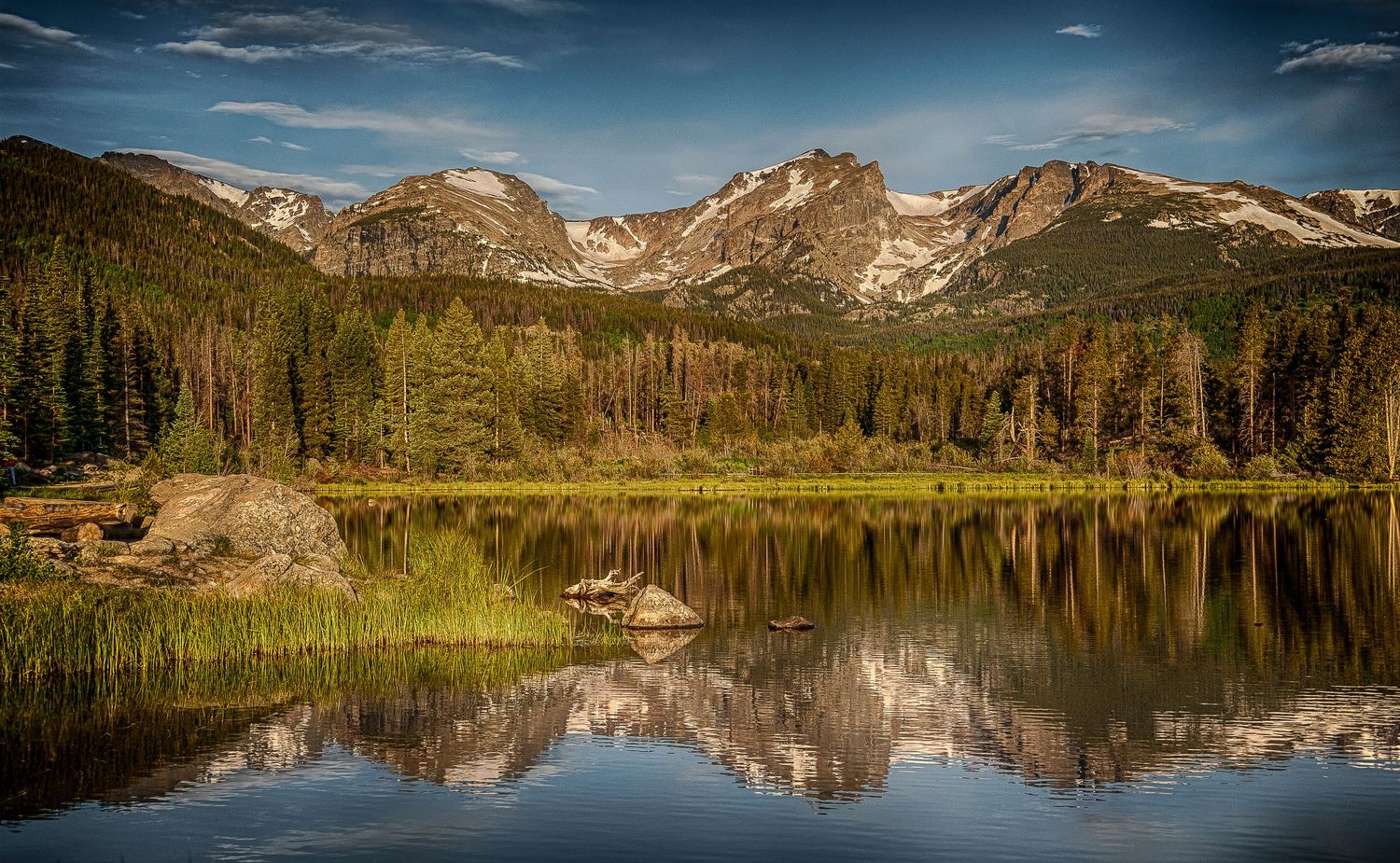 Snow-capped Rocky Mountain peaks reflect perfectly in a calm alpine lake surrounded by evergreen forest.