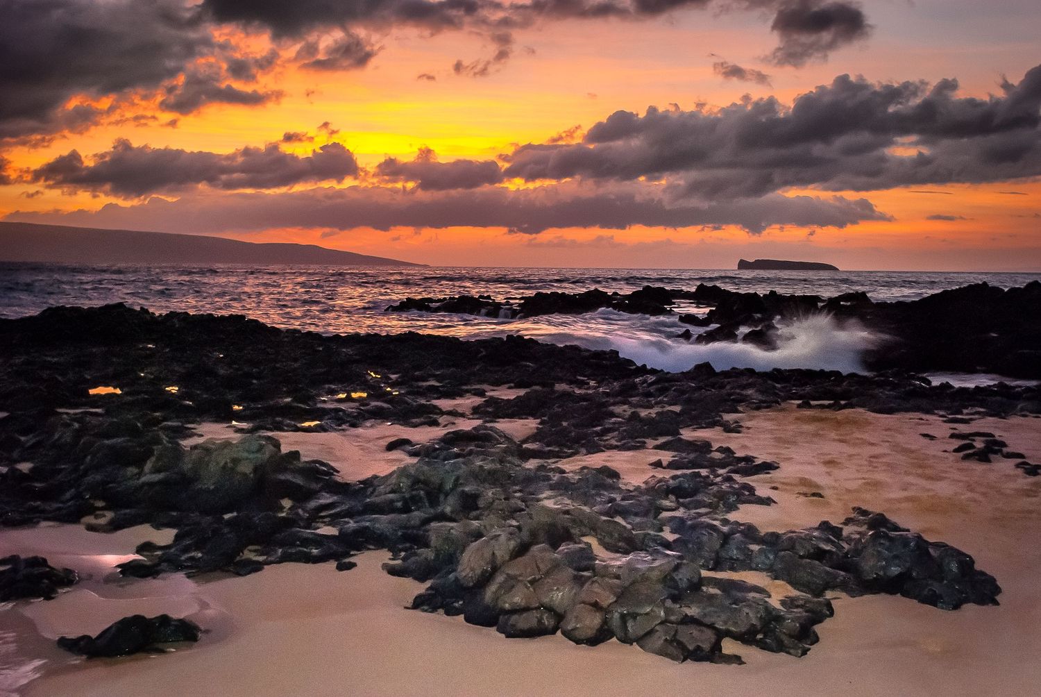 Golden sunset light reflects on rocky black lava shoreline and sandy beach in Hawaii.