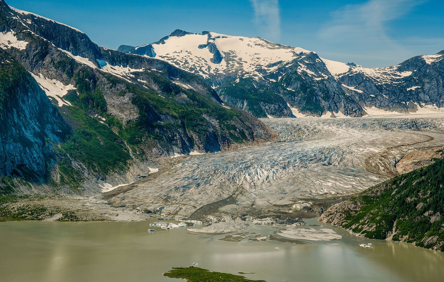 Massive glacier flowing between snow-capped mountains into a turquoise glacial lake in coastal Alaska.