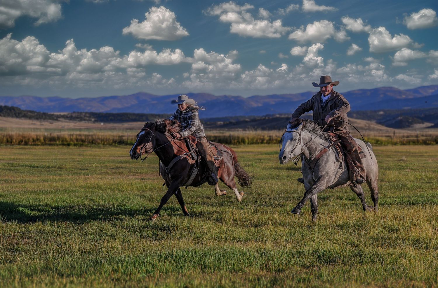 Two cowboys on horseback gallop across a vast green meadow with mountain views.
