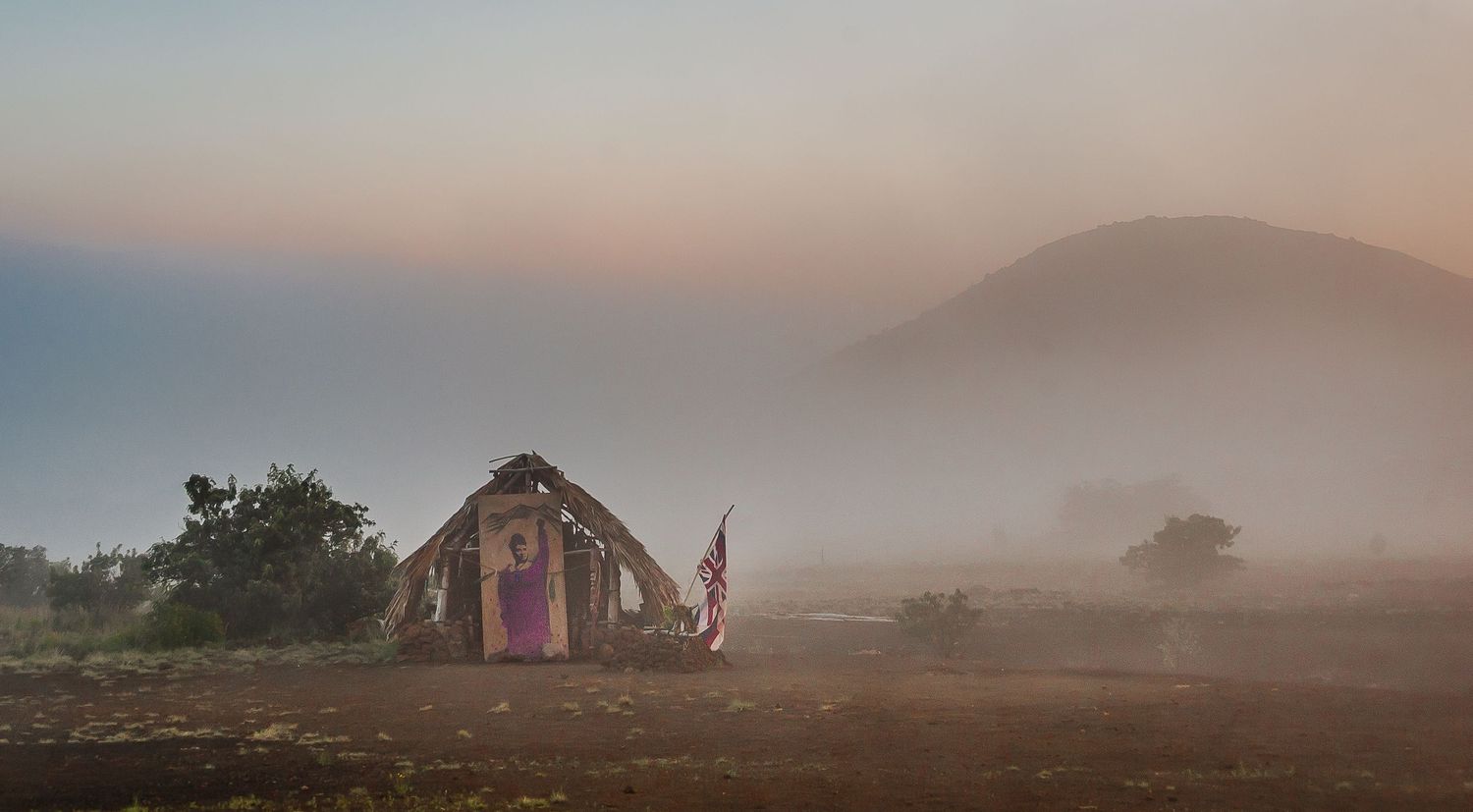 A rustic hut with an American flag stands in misty morning fog with a mountain silhouette at sunrise.