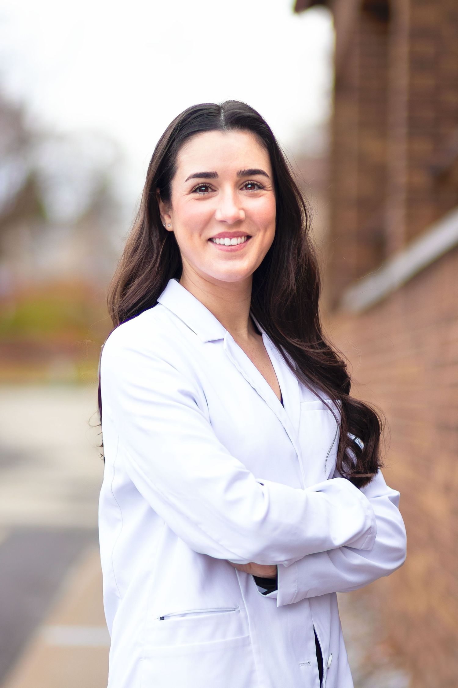 Smiling young female healthcare professional in white lab coat with arms crossed outdoors.