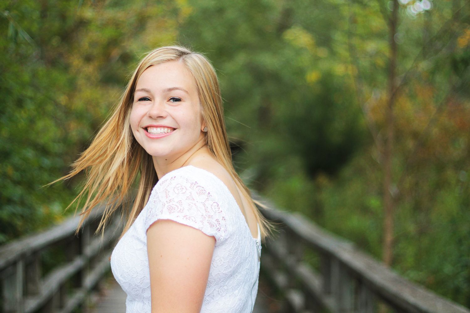 Smiling blonde teenage girl in white lace top on wooden bridge surrounded by green trees.