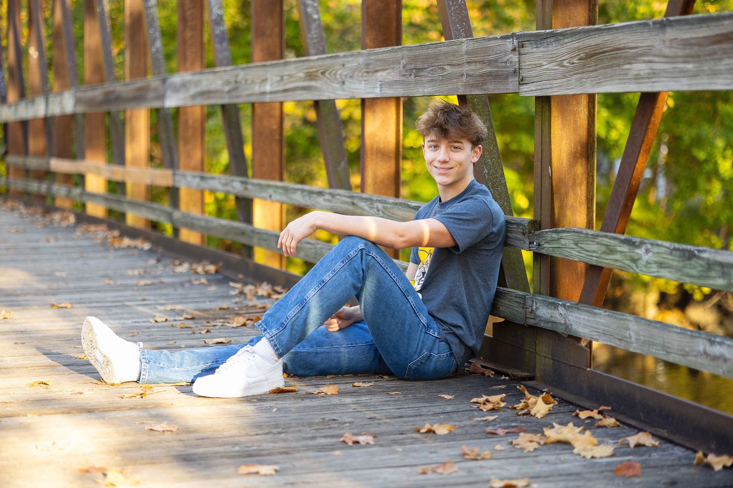 Teenage boy sitting relaxed on wooden bridge deck surrounded by fallen autumn leaves.
