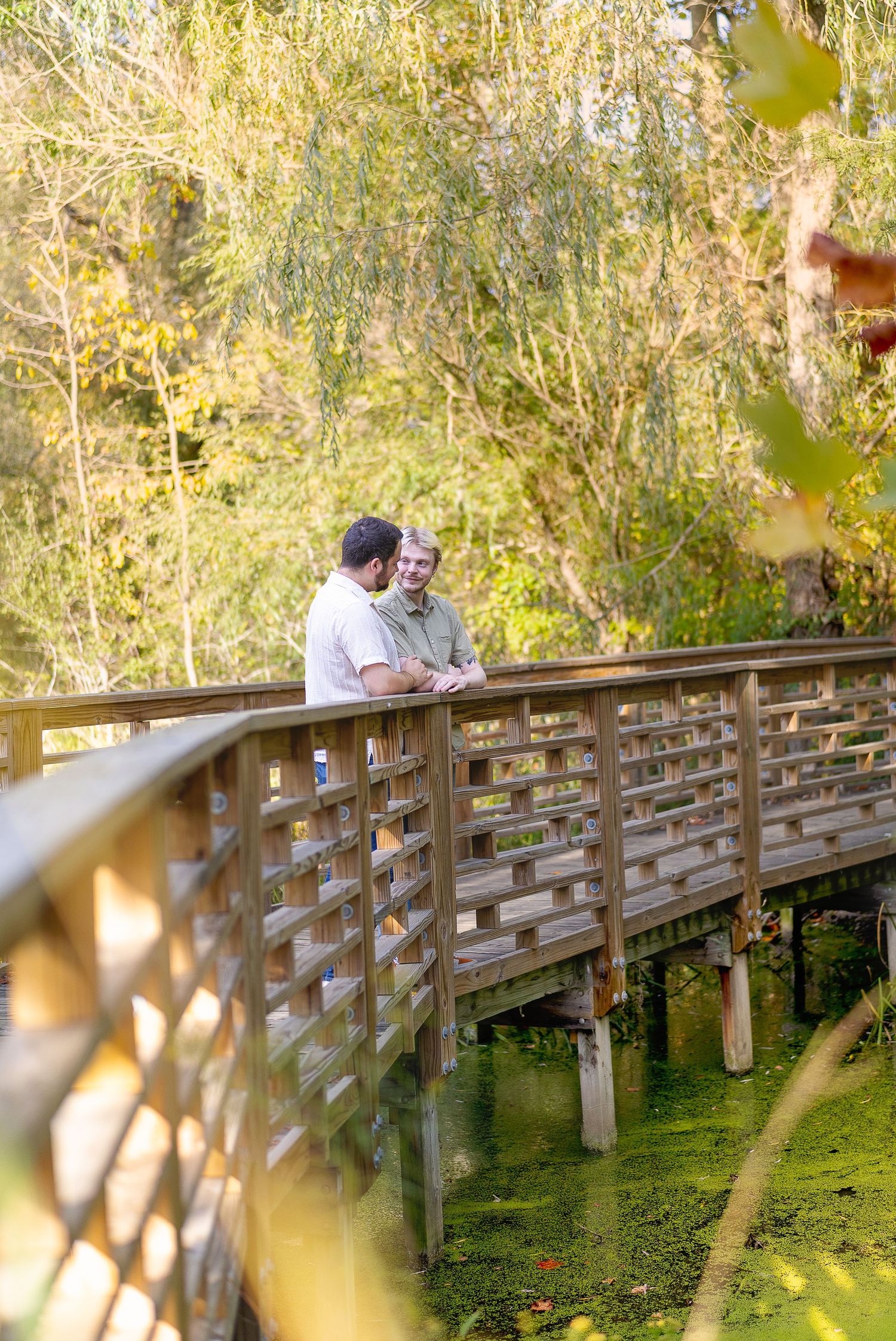 Two men share a joyful moment leaning on a wooden bridge railing surrounded by lush autumn foliage over a green pond.