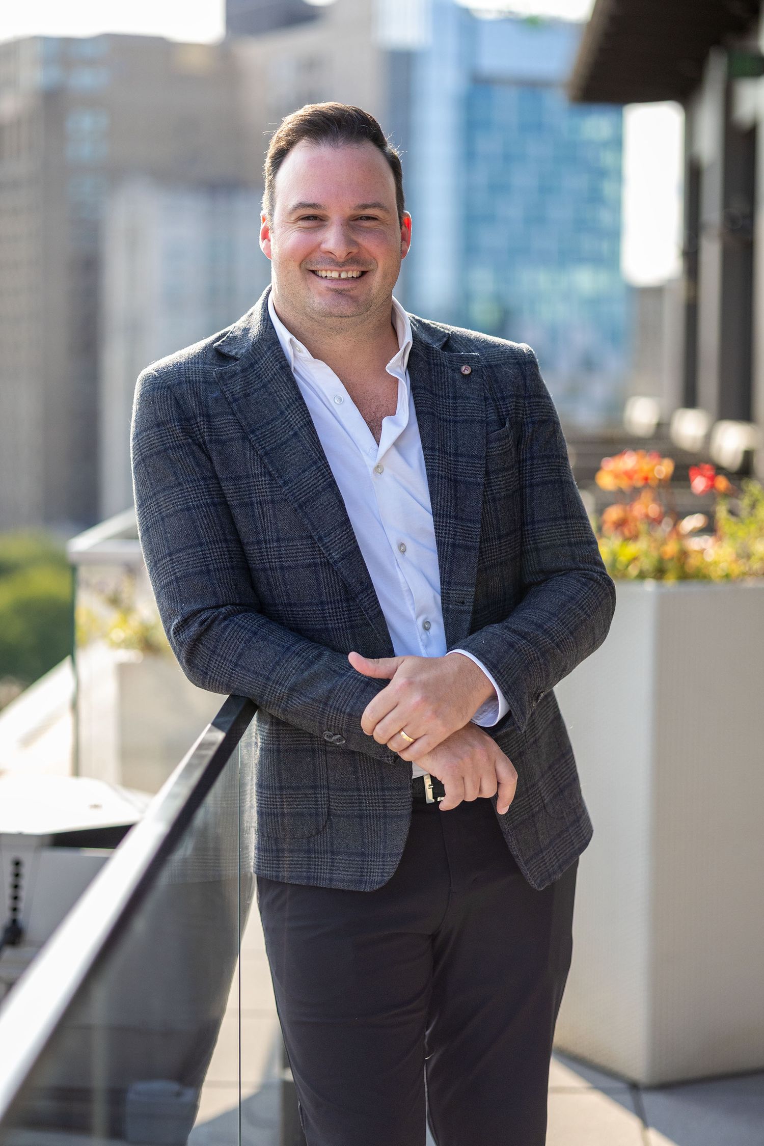 Smiling man in a checkered blazer posing confidently on a rooftop terrace with city buildings behind.