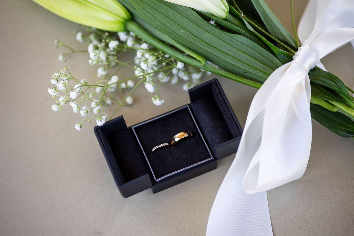 A gold wedding band sits in an open black ring box beside a white floral bouquet with ribbon.