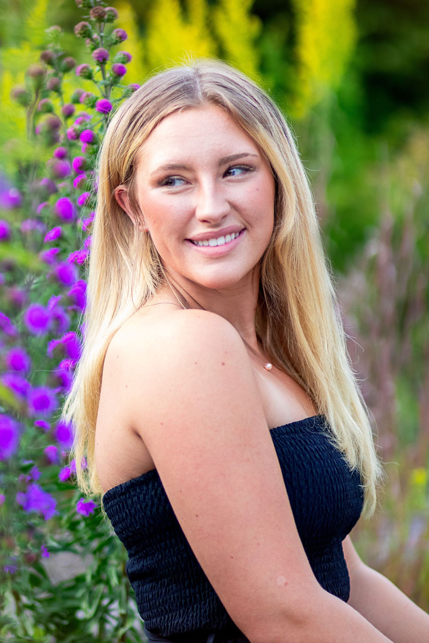 Smiling blonde woman in black tube top posing beside tall purple flowers in garden.