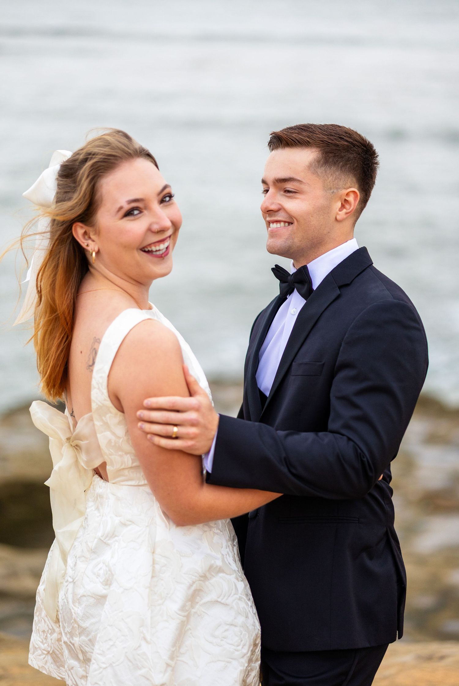Smiling bride in white gown embraced by groom in navy tuxedo near ocean rocks.