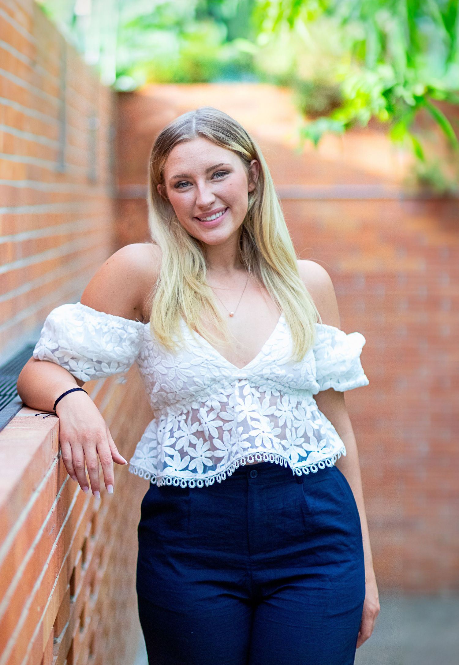 Smiling blonde woman in white off-shoulder lace top leaning against brick wall outdoors.