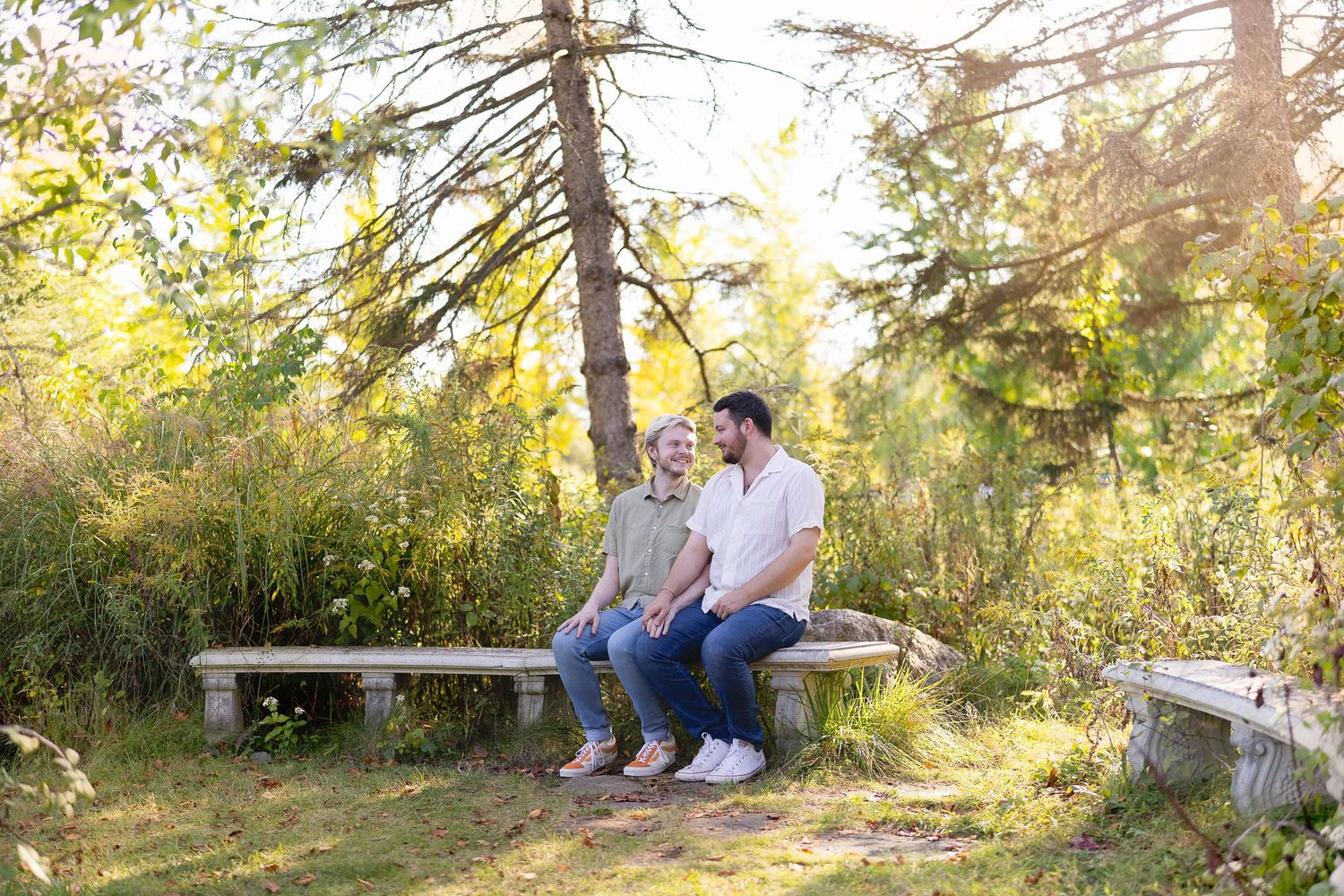 Couple sitting together on a stone bench surrounded by lush green trees in a sunny park.