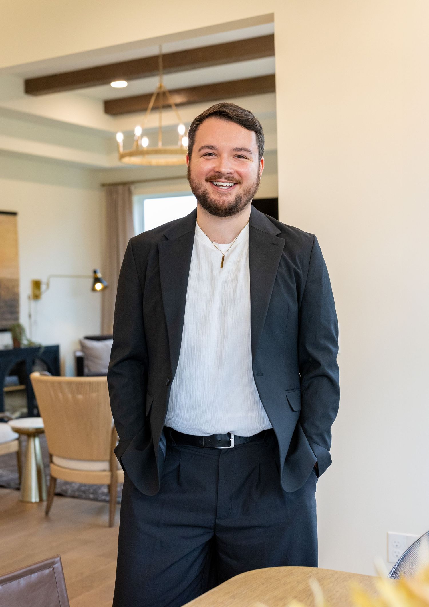 Smiling young man in black blazer and white shirt standing in modern home interior.