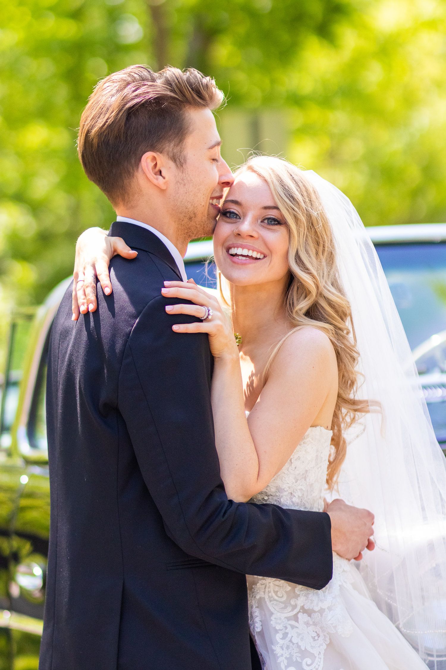 Smiling blonde bride in white lace dress embraces groom in black suit outdoors near a vintage car on wedding day.