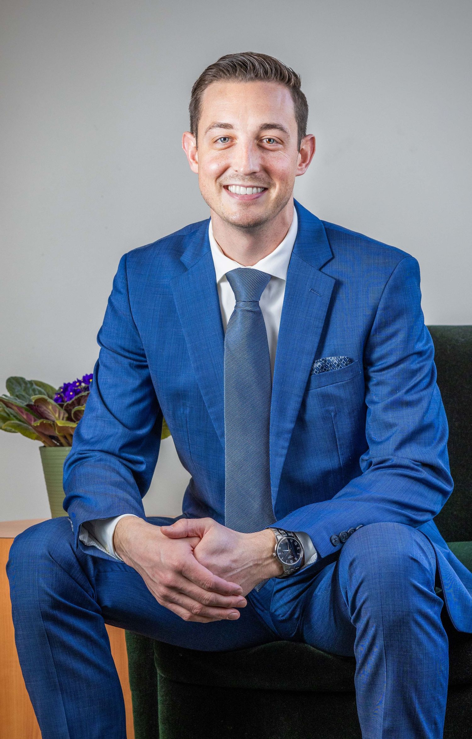 Young professional man in blue suit and gray tie sitting confidently for a business headshot portrait.