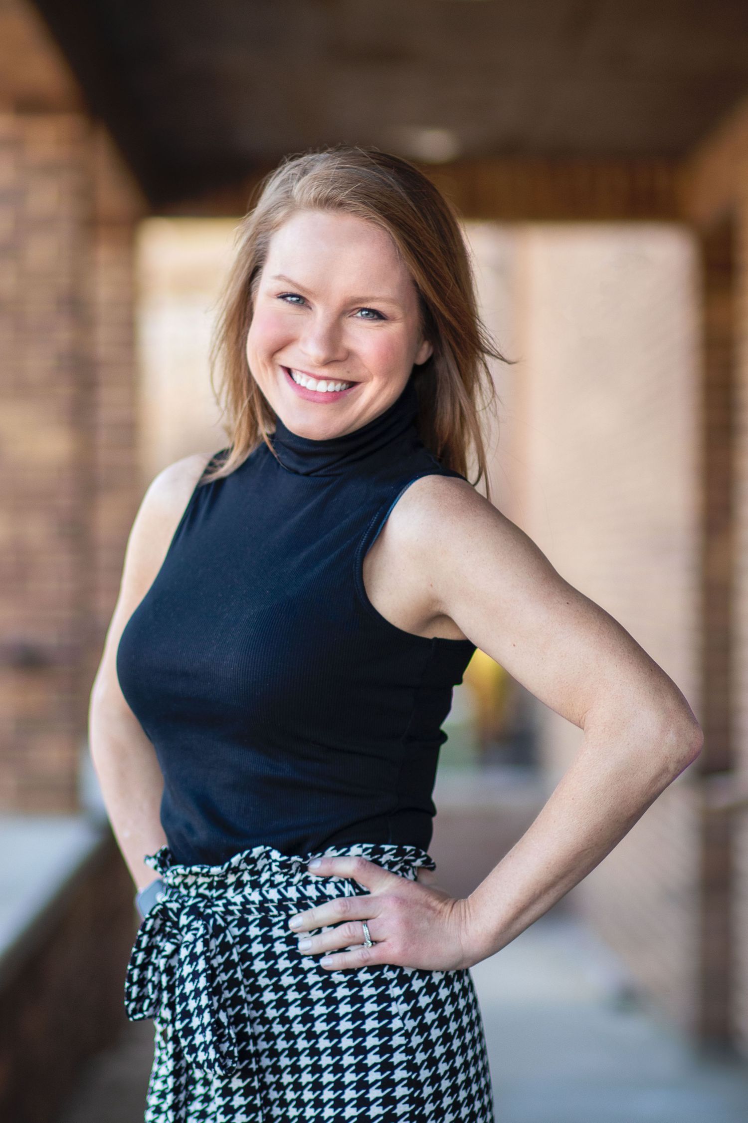 Smiling woman in black turtleneck and houndstooth skirt posing confidently outdoors near brick columns.