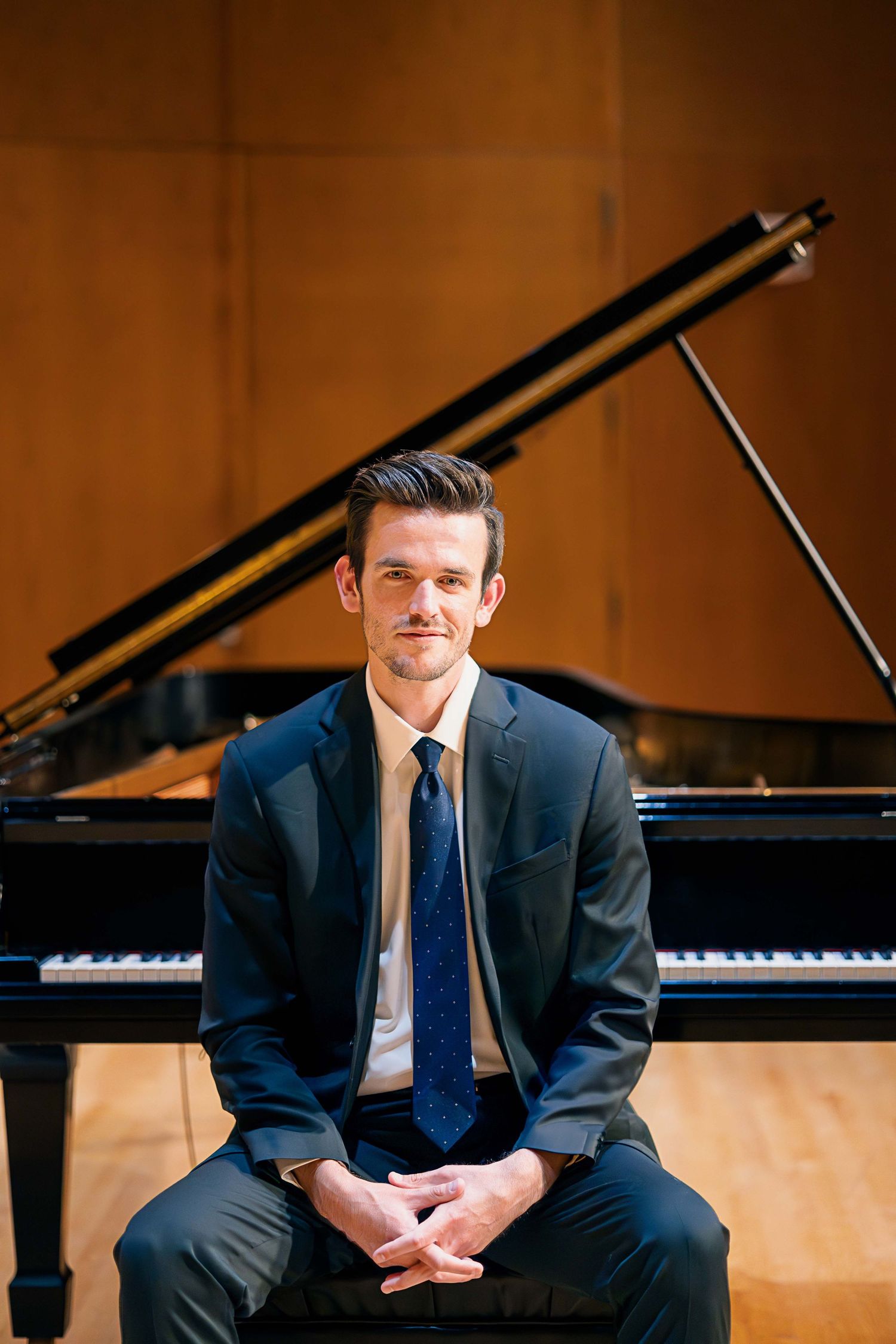 Young male pianist in navy suit seated in front of grand piano on concert hall stage.