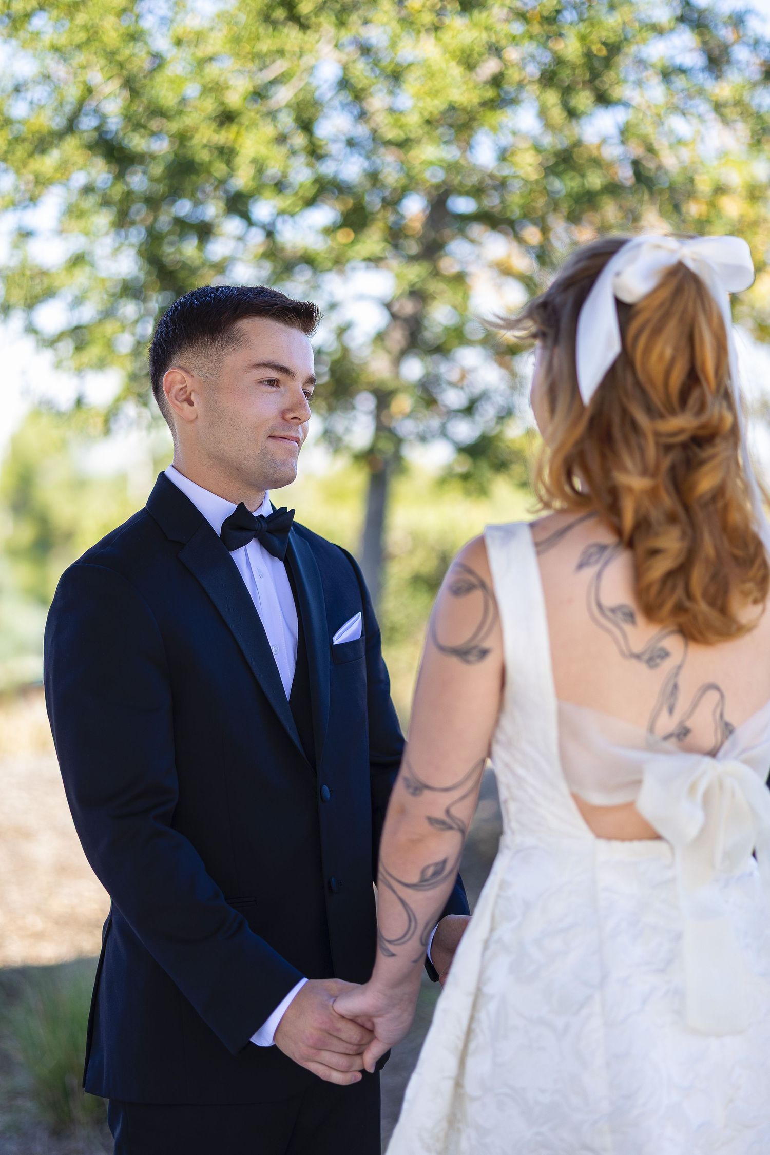A groom in a black tuxedo holds hands with a tattooed bride wearing a white bow in her hair outdoors.