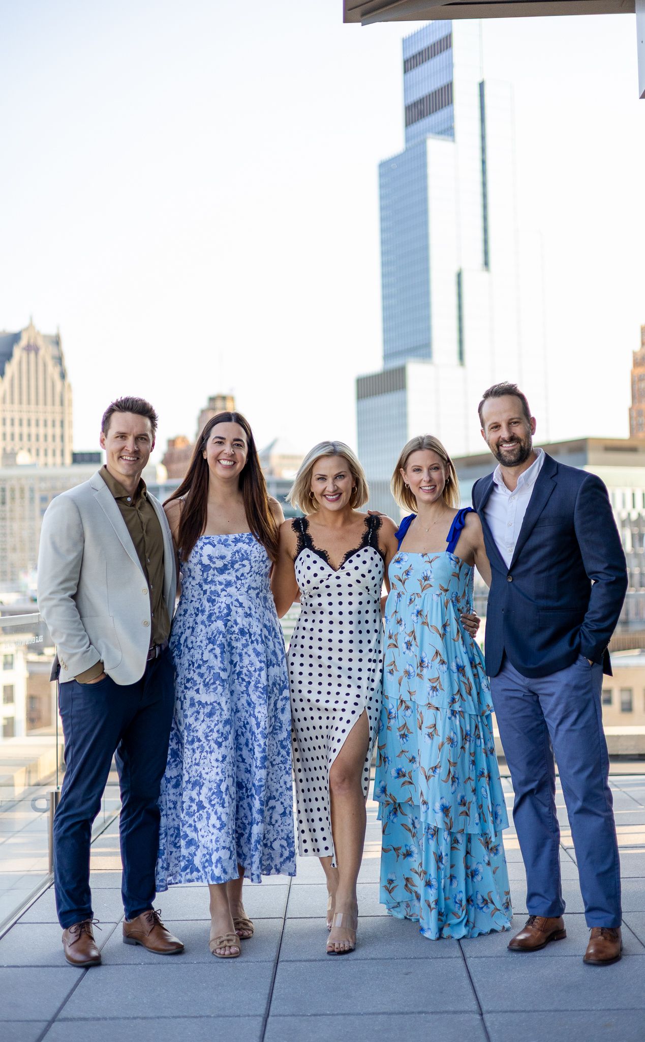 Group of five people smiling together on a rooftop with a city skyline in the background.